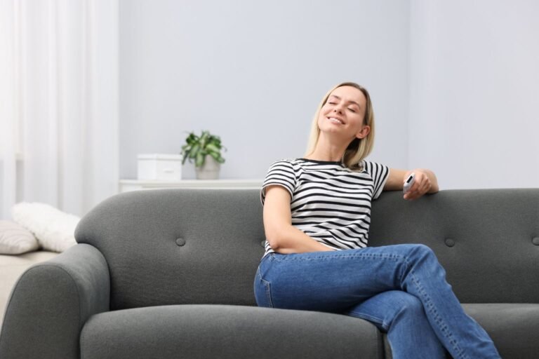 Inicio 13 Uma mulher loira, relaxada e sorrindo com os olhos fechados, desfruta do conforto incomparável em um sofá perfeitamente limpo e convidativo após uma higienização profunda de sofás, segurando um controle remoto.