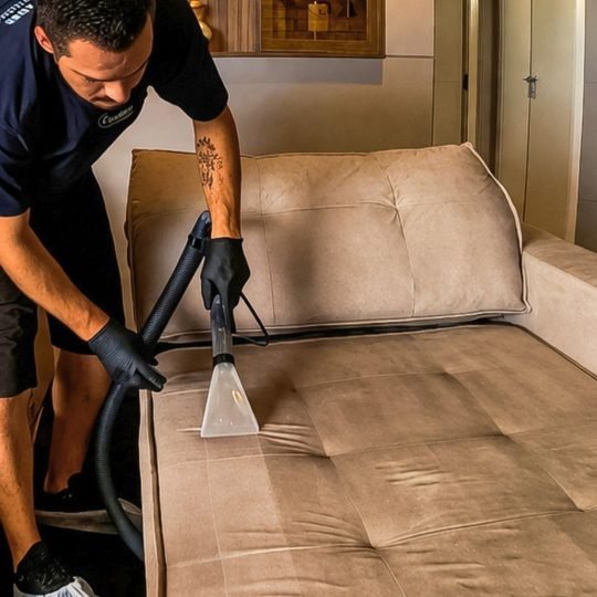 Técnico profissional com luvas pretas e propés nos calçados realizando a limpeza de sofá em Balneário Camboriú. Ele utiliza uma máquina extratora transparente de alta sucção em um sofá retrátil bege, revelando uma nítida faixa limpa que contrasta com a sujeira profunda da área ainda não lavada.