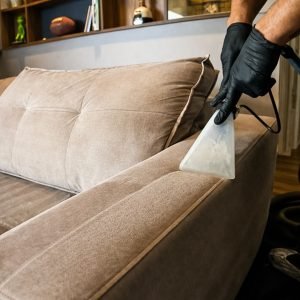 Uma foto em plano aproximado e detalhado foca em um técnico de limpeza profissional. Ele usa luvas de nitrilo pretas e manipula um bocal extrator transparente contra a borda e o apoio de braço de um sofá moderno de tecido bege claro ou taupe. A ferramenta transparente está ativamente extraindo sujeira e umidade, mostrando resultados imediatos em uma linha de tecido visivelmente limpo e húmido. O fundo é um espaço de estar moderno desfocado. Esta imagem ilustra o processo preciso e cuidadoso da limpeza de sofá em Bc.